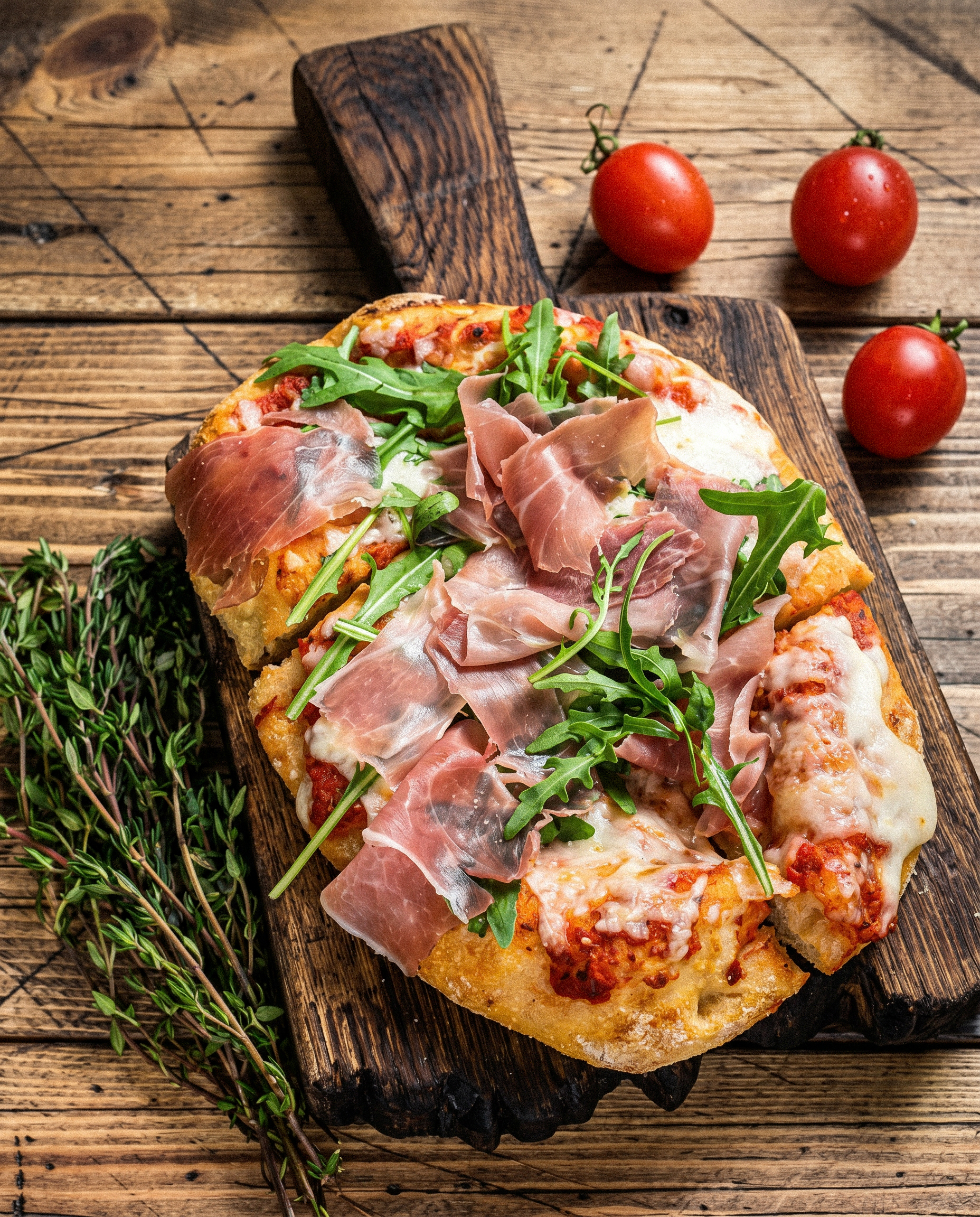 Frisch gebackene Pinsa mit Prosciutto, Rucola, Tomate und Mozzarella auf einem Holzbrett.